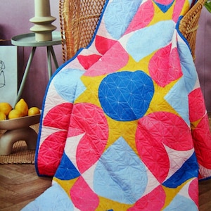 May include: A colorful quilt with a geometric floral pattern in pink, blue, and yellow. The quilt is draped over a wicker chair. The design features large, abstract shapes with a blue border.