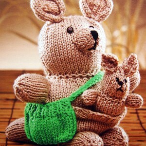 May include: A knitted kangaroo and joey in shades of brown, with a green pouch. The kangaroo has large ears and a small black nose. The joey is nestled in the mother's pouch. The toy is made of yarn.