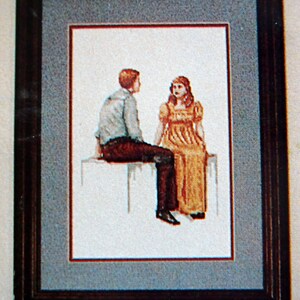 Puede incluir: Una obra de arte de punto de cruz enmarcada que representa a un hombre y una mujer sentados juntos. El hombre lleva una camisa azul claro y pantalones marrones, mientras que la mujer lleva un vestido amarillo. La obra de arte est&aacute; sobre un fondo blanco.
