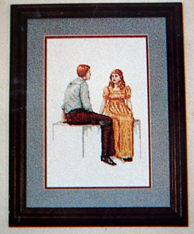 Puede incluir: Una obra de arte de punto de cruz enmarcada que representa a un hombre y una mujer sentados juntos. El hombre lleva una camisa azul claro y pantalones marrones, mientras que la mujer lleva un vestido amarillo. La obra de arte est&aacute; sobre un fondo blanco.