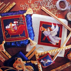 May include: A collection of Christmas-themed needlepoint crafts. The image features a square pillow with a Santa's sleigh design, a needlepoint angel, and other small ornaments. Gold tassels, frames, and various craft supplies are also visible.