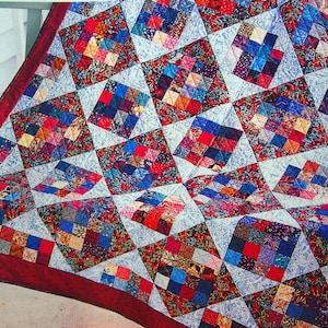 Puede incluir: Una colcha de patchwork con un patrón repetitivo de cuadrados multicolores en tonos rojos, azules, amarillos y morados, intercalados con bloques de tela estampada en azul claro. La colcha tiene un borde de tela rojo oscuro sólido.