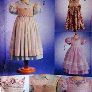 May include: A sewing pattern for three different styles of girls' dresses. The first dress is white with a blue collar and sleeves, the second dress is pink and white gingham, and the third dress is floral print.