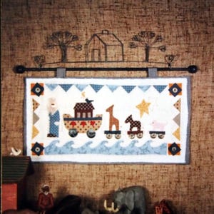 May include: A colorful quilted wall hanging depicting a whimsical scene of Noah's Ark with animals on a blue and white wave background. The ark is brown with red hearts and a bird on top. The animals include a giraffe, a horse, and a pig. The hanging is framed with a blue and white patterned border.