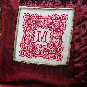 May include: A claret velvet cushion with a stitched design. The centre has a square of off-white fabric with a red embroidered design and the letter "M". The cushion has a gold trim.