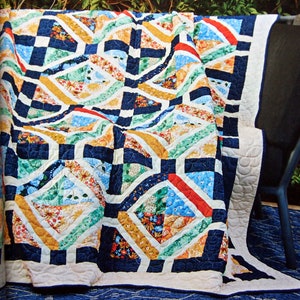Op de afbeelding: Een patchwork quilt met een herhalend geometrisch patroon van ruiten en vierkanten. Het ontwerp bevat marineblauwe tussenstroken met diverse bloemrijke en bedrukte stoffen blokken in tinten geel, rood, groen en lichtblauw, afgewerkt met een crèmekleurige rand.