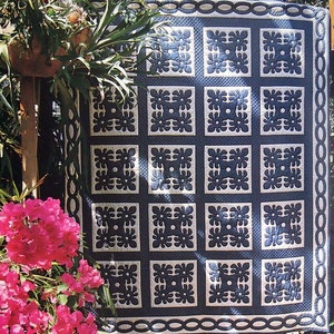 May include: A blue and white quilt with a repeating pattern of stylised flowers. The quilt is hanging on a wooden frame and is surrounded by greenery.