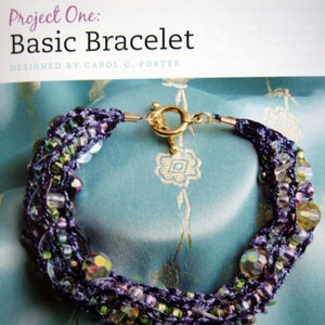 May include: A multi-strand bracelet with purple, green, and clear beads, and a gold clasp. The bracelet is on a blue fabric background.