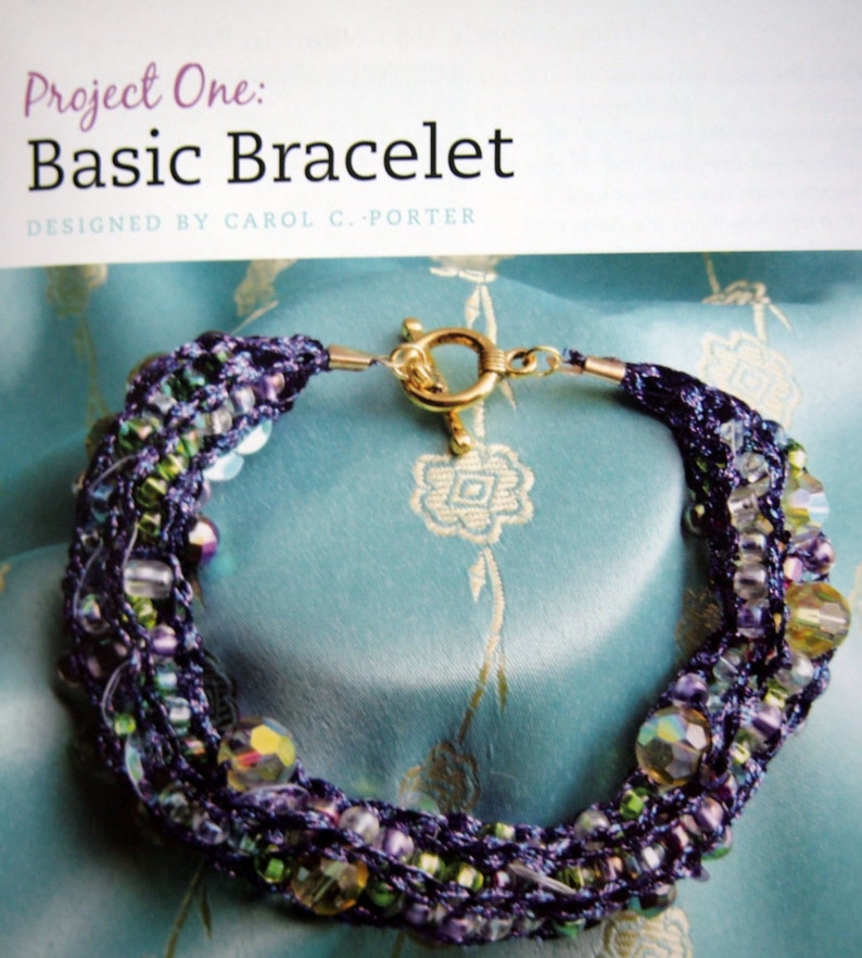 May include: A multi-strand bracelet with purple, green, and clear beads, and a gold clasp. The bracelet is on a blue fabric background.