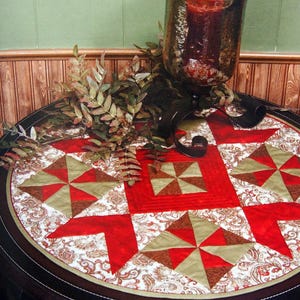 May include: A round table with a quilted table topper featuring a red and tan starburst pattern. A large glass candle holder with a red candle sits on the table, along with decorative greenery and a black metal accent.