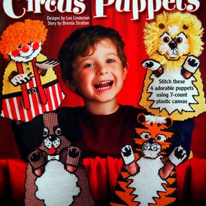 May include: A craft book cover featuring four colorful puppets: a clown, a dog, a tiger, and a lion. The puppets are made with plastic canvas and are designed to be stitched. The book cover also includes the title "Circus Puppets" and the text "Stitch these 4 adorable puppets using 7-count plastic canvas."