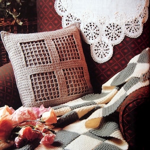 May include: A crocheted pillow with a square grid design, a checkered blanket, and a white lace doily rest on a patterned armchair. A bouquet of pink roses adds a touch of color to the scene. The blanket has a fringe.