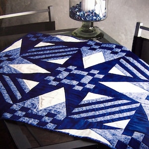 Könnte beinhalten: Eine blaue und weiße Steppdecke mit geometrischem Muster. Die Steppdecke besteht aus Quadraten und Dreiecken und hat einen weißen Hintergrund mit blauen Akzenten. Die Steppdecke wird auf einem Tisch mit Glasplatte gezeigt.