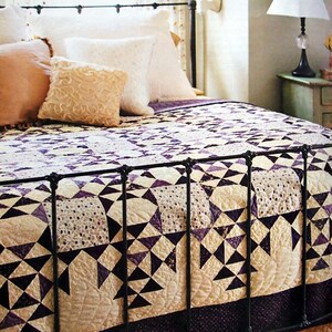 Puede incluir: Una cama con un marco de metal negro y una colcha blanca y morada. La colcha tiene un patrón geométrico de triángulos y cuadrados. La cama tiene una cabecera blanca y un piecero blanco. Hay almohadas en la cama, incluyendo una almohada de color canela, una almohada blanca y una almohada crema con un diseño de encaje.