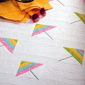 May include: A white quilt patterned with colourful kites. The kites have yellow, green, blue, and pink stripes. A yellow towel, red sunglasses, and a green handbag are in the upper left corner.