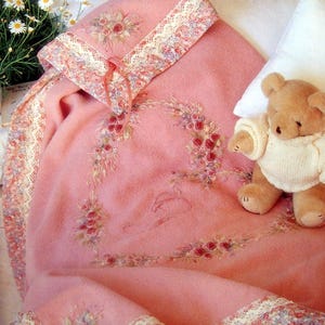 Peut inclure: Une couverture rose pour bébé avec une bordure en dentelle blanche et des motifs floraux brodés. Un ours en peluche est assis sur la couverture.
