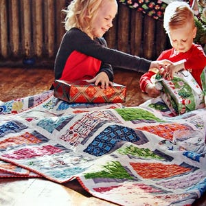Op de afbeelding: Twee kinderen zitten op een kleurrijk patchwork-deken en openen cadeaus. Het meisje draagt een rood shirt en een zwarte trui, en de jongen draagt een rood shirt. Het deken heeft verschillende patronen, waaronder bloemen, geometrische vormen en sleutels.
