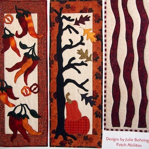 May include: Three fabric wall hangings. The first features red and orange chili peppers with green stems and pretzel shapes on a cream background. The second has a black tree, leaves, and a pumpkin on a cream background. The third has burgundy wavy stripes.