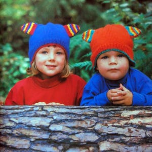 Op de afbeelding: Twee kinderen dragen kleurrijke gebreide mutsen met oorflappen. De mutsen zijn blauw en rood met gele en witte strepen. De kinderen zitten voor een boomstam.