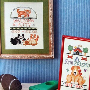 Puede incluir: Dos dise&ntilde;os de bordado de punto de cruz enmarcados. El primer dise&ntilde;o presenta un gato con el texto "Welcome Kitty Ginger June 2003". El segundo dise&ntilde;o presenta un perro con el texto "A New Friend Simon 2005".