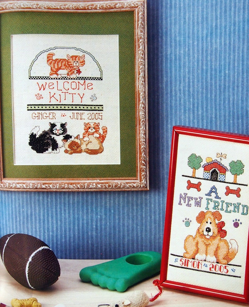 Puede incluir: Dos dise&ntilde;os de bordado de punto de cruz enmarcados. El primer dise&ntilde;o presenta un gato con el texto "Welcome Kitty Ginger June 2003". El segundo dise&ntilde;o presenta un perro con el texto "A New Friend Simon 2005".