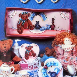May include: A set of three painted wooden heart-shaped boxes with teddy bear designs. The boxes are decorated with pearls, ribbons, and lace. The largest box has a teddy bear holding a blue ribbon and the text "Say it with a smile". The middle box has a teddy bear holding a blue ribbon and the text "The sweetest of them all". The smallest box has a teddy bear holding a blue ribbon and the text "With love and a smile".