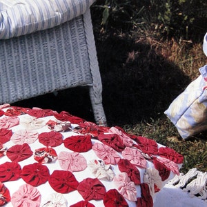 May include: A white wicker chair with a blue and white striped cushion. A white pillow with red and pink fabric rosettes is on the ground in front of the chair.