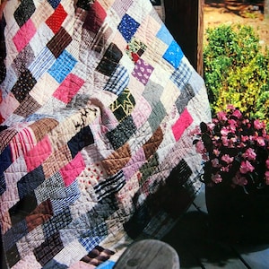 May include: A patchwork quilt made of various fabrics in shades of pink, blue, brown, and white. The quilt is folded and draped over a wooden chair, with a portion of the quilt hanging off the chair.
