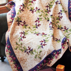 May include: A beige quilt featuring a repeating pattern of green, purple, and red geometric shapes. The quilt has a dark blue border with a floral design in red and purple. The quilt is draped over a dark green sofa.