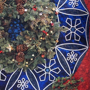 May include: A festive Christmas tree skirt featuring a vibrant blue snowflake pattern with white accents. The skirt is adorned with a lush evergreen wreath, pinecones, and twinkling lights, creating a warm holiday ambiance.