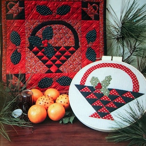 Op de afbeelding: Een rode en zwarte gewatteerde wandhanger en een kerst-borduurring. De wandhanger en de ring tonen een mandontwerp met hulstbladeren en bessen. Sinaasappels en groen maken de feestelijke scène compleet, wat een kerstsfeer creëert.