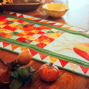 Peut inclure: Un chemin de table en patchwork coloré avec une bordure verte et une applique en tissu blanc représentant du maïs. Le chemin de table est posé sur une table en bois avec une citrouille et d'autres décorations d'automne.