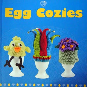 May include: Egg cozies in various designs, including a chick, a jester's hat, and a flower pot. The cozies are knitted and sit atop white egg cups. The background is blue with the words "Egg Cozies" in yellow.