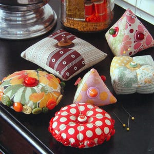 May include: A selection of handmade pincushions in diverse shapes and patterns. The pincushions display floral, polka dot, and striped fabrics, embellished with buttons and decorative elements. Colours include red, pink, green, yellow, and brown.