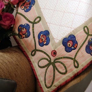Peut inclure: Un coussin matelassé avec un centre en tissu blanc à motif quadrillé rouge, bordé d'un tissu à rayures roses et blanches. La bordure est décorée de vignes en tissu vert et de fleurs en tissu bleu avec des cœurs orange.