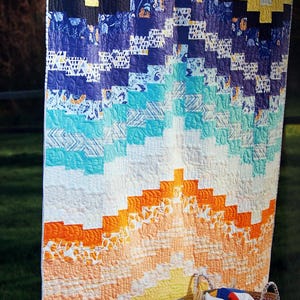 Op de afbeelding: Een kleurrijk patchwork-deken met een chevron-patroon in tinten blauw, turquoise, oranje en geel. Het deken hangt aan een waslijn en is omgeven door groen gras.