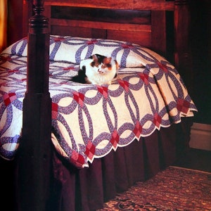 May include: A calico cat rests on a vintage double wedding ring quilt. The quilt features a white background with red and blue interlocking rings and diamond accents. The bed has a dark wood frame and a burgundy bed skirt.