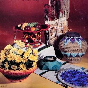 May include: A collection of woven baskets in various sizes and colors, including a fruit bowl with red, green, and yellow fruit, a large brown basket with blue and white accents, a smaller blue and green basket, and a round blue and purple coaster. The baskets are arranged on a white and brown rug with a southwestern pattern.