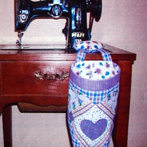 May include: A vintage black sewing machine with a purple and white fabric hanging basket with a heart design. The sewing machine is on a wooden table.