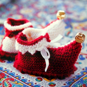 May include: Red and white knitted baby booties with a white ribbon and gold bells. The booties are on a colorful patterned fabric.