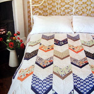 May include: A white bed with a white and blue chevron patterned quilt. The bed has a white headboard with a decorative metal design. The bed is in a room with a fireplace and a floral wallpaper.