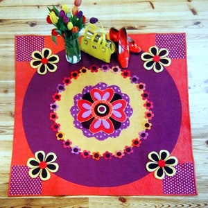 May include: A purple and orange round rug with a large flower design in the center. The rug has a white polka dot border and is decorated with smaller flowers around the edge. There are two red shoes and a yellow purse on the rug.