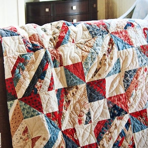 Peut inclure: Une courtepointe patchwork rouge, blanche et bleue avec un motif matelassé. La courtepointe est faite de différents tissus avec des motifs différents et est pliée sur un canapé.