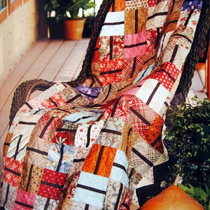 May include: A patchwork quilt with brown, red, orange, and blue fabric squares, draped over a wicker chair on a porch. A large gourd sits on the floor next to the chair.