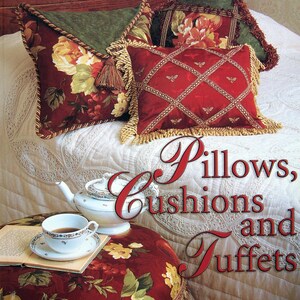 May include: A book titled Pillows, Cushions and Tuffets by Carol Zentgraf featuring a cover image of decorative pillows with floral and lattice patterns, a white teapot, a teacup on a saucer, and an open book resting on a floral upholstered tuffet.