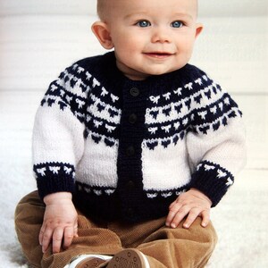 May include: A baby wearing a white and navy blue knitted cardigan with a traditional Scandinavian pattern. The cardigan has a button closure and the baby is wearing brown corduroy pants.