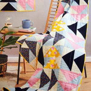 Op de afbeelding: Een kleurrijke quilt met een geometrisch patroon van zwarte, roze, gele en blauwe driehoeken. De quilt is over een rieten stoel gedrapeerd, met een bijpassende wandophanging. De quilt heeft een gele rand.