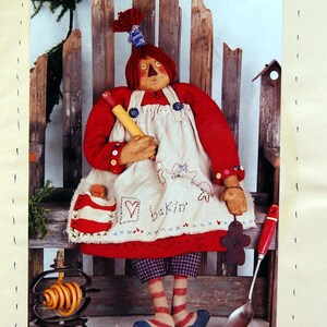 Peut inclure: Poupée d'art populaire nommée "Cookie" portant un haut rouge et un tablier, tenant un rouleau à pâtisserie. Le tablier porte un cœur et le mot "bakin'" brodé. La poupée mesure 76 cm et a un bonhomme en pain d'épices et une cuillère.