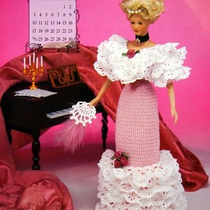 May include: A doll wearing a pink and white crocheted dress with a ruffled skirt and a white ruffled collar. The doll has blonde hair and is holding a white crocheted accessory. A calendar for March 1996 is visible.