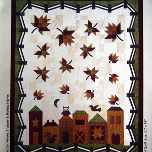Puede incluir: Un patrón de colcha con una ciudad con casas, un granero, una estrella, una calabaza y un búho. Las casas están rodeadas de hojas que caen en tonos de marrón, rojo y verde. La colcha está enmarcada por una valla verde y negra. El texto "Hometown Harvest" está escrito en marrón en la parte superior de la colcha.